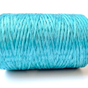 May include: A spool of light blue raffia ribbon. The ribbon is tightly wound into a cylindrical shape. The color is a soft, muted blue, and the texture appears slightly rough. Ideal for crafting and gift wrapping.