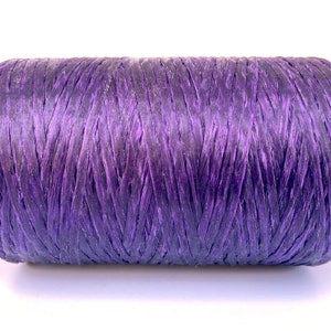 May include: A cylindrical spool of purple raffia ribbon. The ribbon is tightly wound, displaying a woven texture. The colour transitions from a lighter purple to a darker shade, creating a gradient effect. Suitable for crafting and gift wrapping.