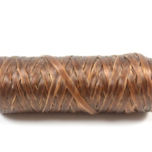 May include: A spool of brown raffia ribbon, tightly wound and ready for crafting. The ribbon has a woven texture and a natural, earthy tone, perfect for adding a rustic touch to various projects. The spool is set against a plain white background.