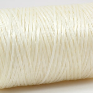 May include: A spool of off-white raffia ribbon. The ribbon is tightly wound around a cardboard core, creating a cylindrical shape. The raffia strands have a slightly textured appearance, and the overall color is a soft, creamy white.
