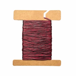 May include: A spool of burgundy-colored raffia ribbon. The ribbon is tightly wound around a cardboard spool with a slot at the top. The raffia has a textured appearance, and the color varies slightly throughout.