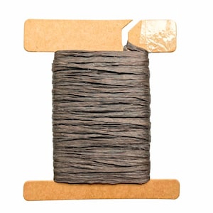 May include: A spool of dark brown raffia ribbon. The ribbon is tightly wound around a cardboard spool. The spool has a rectangular shape with a small tab at the top. The ribbon appears to be made of natural fibers.