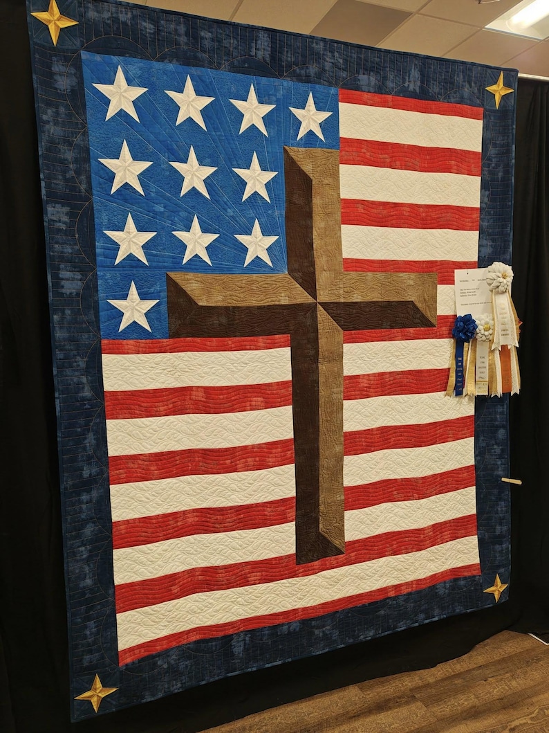 Pode incluir: Uma grande tape&ccedil;aria de parede acolchoada com o design da bandeira americana e uma cruz marrom. A bandeira tem listras vermelhas e brancas, um campo azul com estrelas brancas e uma borda azul escura com estrelas douradas.
