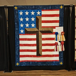 Pode incluir: Um edredom patri&oacute;tico com a bandeira americana e uma cruz marrom sobreposta. O edredom tem listras vermelhas e brancas, estrelas azuis e uma borda azul com estrelas douradas. As fitas de pr&ecirc;mio est&atilde;o anexadas.