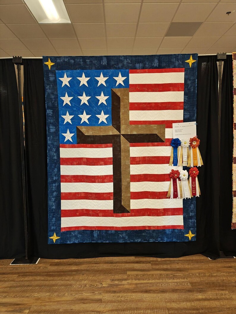 Pode incluir: Um edredom patri&oacute;tico com a bandeira americana e uma cruz marrom sobreposta. O edredom tem listras vermelhas e brancas, estrelas azuis e uma borda azul com estrelas douradas. As fitas de pr&ecirc;mio est&atilde;o anexadas.