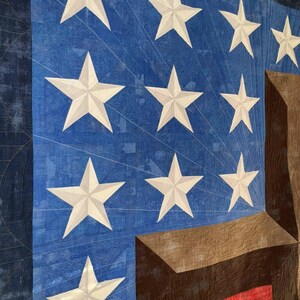 Pode incluir: Um close-up de um edredom com um campo azul com estrelas brancas e uma se&ccedil;&atilde;o listrada marrom e vermelha. O edredom tem um tema patri&oacute;tico, com o design de estrelas e listras.