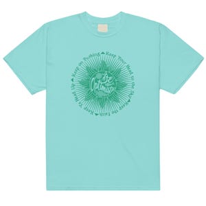 May include: A pale turquoise t-shirt with a green circular design. The design includes the words "Be Optimistic" and surrounding text that reads "Keep Ya Head Up, Keep on Pushing, Keep Your Head to the Sky, Keep the Faith."