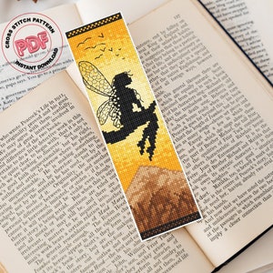 May include: A cross stitch bookmark featuring a silhouette of a fairy with wings sitting on a mountain. The background is a sunset with a gradient of orange, yellow, and brown. The bookmark is surrounded by a white border.