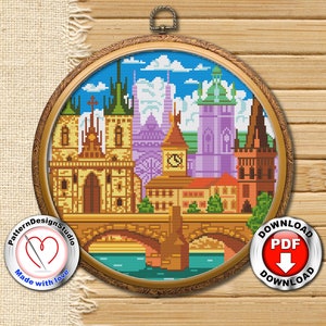May include: A colorful cross-stitch pattern of the Prague skyline, featuring a bridge, buildings, and a river. The pattern is framed in a gold-colored hoop and the words "Prague" and "Download PDF" are visible below the hoop.