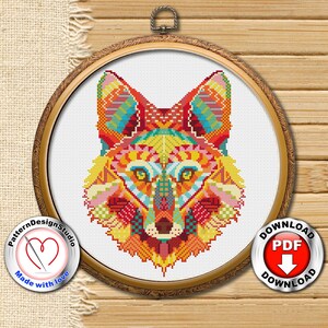 May include: A colourful cross-stitch pattern of a fox's head in a wooden embroidery hoop. The fox is depicted in a geometric style with vibrant colours, including red, orange, yellow, green, blue, and purple. The pattern is framed by a wooden hoop with a gold finish. The words "Pattern Design Studio" and "Made with love" are printed on a red heart-shaped label below the hoop. The words "Download PDF" are printed on a red circle with a downward arrow.