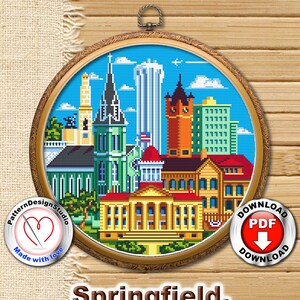 May include: Cross-stitch pattern of the Springfield, Illinois skyline. The pattern features a variety of buildings, including the state capitol building, a church, and a skyscraper. The pattern is in a circular frame with a gold border.
