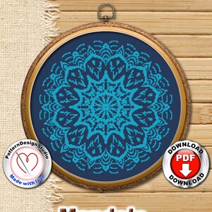 May include: A blue and teal mandala cross stitch pattern in a wooden embroidery hoop. The pattern is framed with a dark blue background. The text "Mandala" is printed below the hoop. A red heart with a needle is in the lower left corner of the image. A red circle with the text "Download PDF" is in the upper right corner of the image.