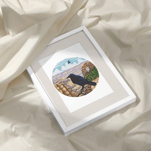 May include: A framed print of a black raven perched on a cliff overlooking a canyon. The raven is in the foreground, with the canyon in the background. The print is in a white frame with a beige mat.