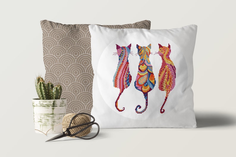 Pode incluir: Tr&ecirc;s gatos coloridos e com padr&otilde;es s&atilde;o representados em um design circular em uma almofada branca. Os gatos est&atilde;o de costas para o espectador, com suas caudas enroladas ao redor de seus corpos. A almofada &eacute; um acento decorativo para uma casa.