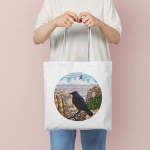 May include: White tote bag with a black crow perched on a rock overlooking a canyon. The design is in a circle with a blue sky and white clouds in the background.