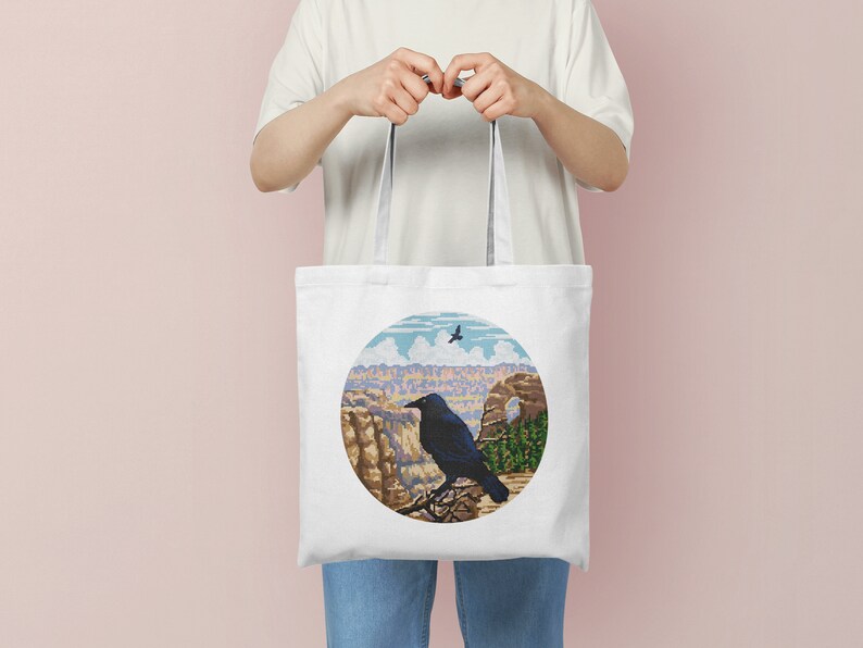 May include: White tote bag with a black crow perched on a rock overlooking a canyon. The design is in a circle with a blue sky and white clouds in the background.