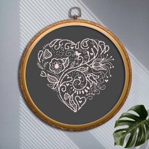 Peut inclure: Un cercle de broderie doré avec un fond gris foncé et un motif de cœur floral blanc. Le cœur est composé de volutes, de feuilles et de fleurs.