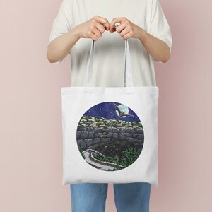 May include: White tote bag with a circular design featuring a night sky with a crescent moon, stars, and a bird flying over a stone wall.