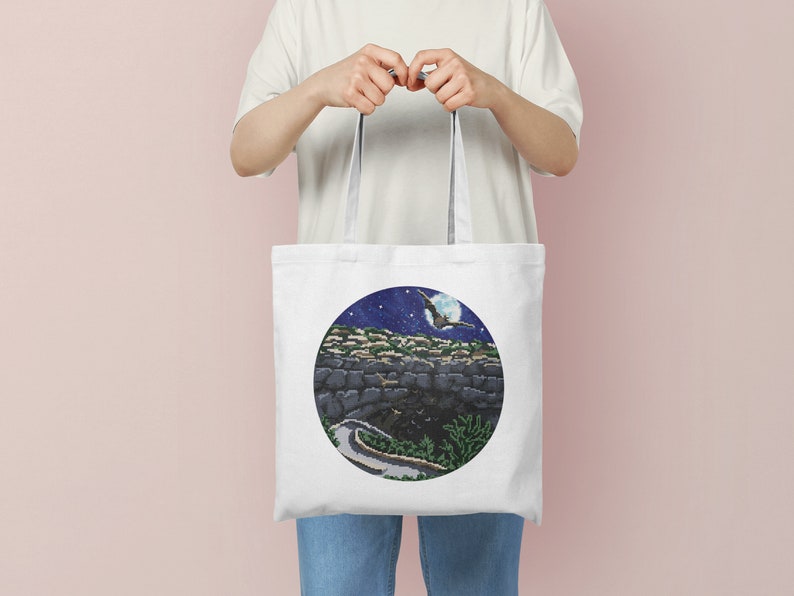 May include: White tote bag with a circular design featuring a night sky with a crescent moon, stars, and a bird flying over a stone wall.