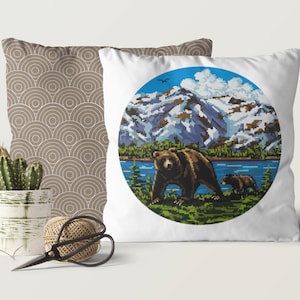 Pu&ograve; includere: Cuscino bianco con un design circolare che presenta un orso bruno e un cucciolo che camminano in un paesaggio montano con un lago. Lo sfondo include un cielo blu con nuvole bianche e montagne innevate.