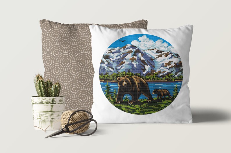 Pu&ograve; includere: Cuscino bianco con un design circolare che presenta un orso bruno e un cucciolo che camminano in un paesaggio montano con un lago. Lo sfondo include un cielo blu con nuvole bianche e montagne innevate.