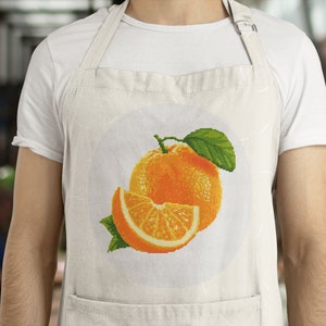 May include: A white apron with a pocket and an orange fruit design. The design features a whole orange and a slice of orange with green leaves.