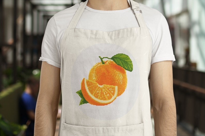 May include: A white apron with a pocket and an orange fruit design. The design features a whole orange and a slice of orange with green leaves.
