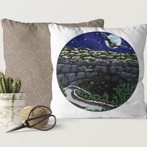 May include: A white pillow with a circular design featuring a night sky with stars and a full moon. A bat flies in front of the moon. The scene is set against a dark, rocky landscape with a winding path.