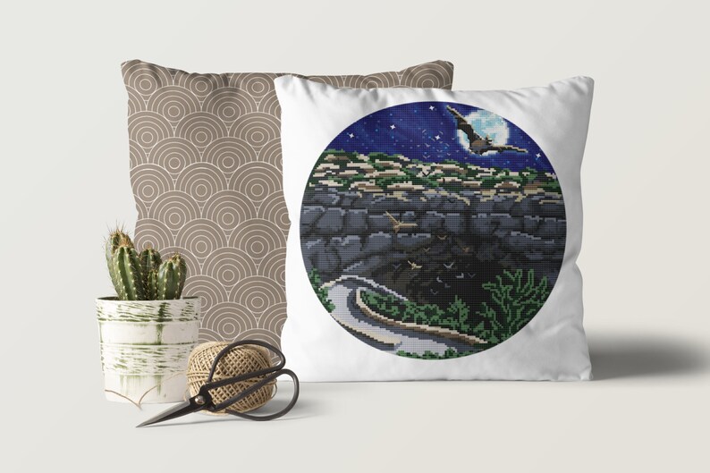 May include: A white pillow with a circular design featuring a night sky with stars and a full moon. A bat flies in front of the moon. The scene is set against a dark, rocky landscape with a winding path.
