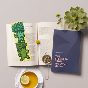 May include: A book with a bookmark featuring a green broccoli character. The book is titled "The Speckled Band and Other Stories". A cup of tea with a lemon slice is in the foreground.