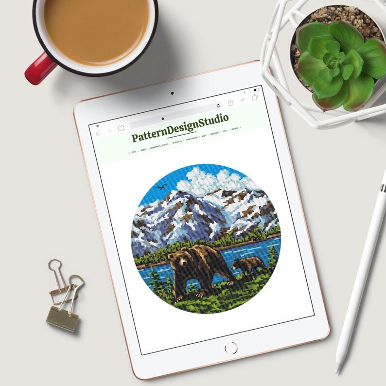 Pu&ograve; includere: Un'illustrazione digitale di un orso bruno e un cucciolo che camminano attraverso un paesaggio montano con un lago e cime innevate. L'illustrazione &egrave; in una cornice circolare e si trova su un sito web chiamato PatternDesignStudio.