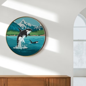 Pode incluir: Uma impress&atilde;o redonda emoldurada de uma orca preta e branca saltando para fora da &aacute;gua em frente a uma cordilheira. A &aacute;gua &eacute; de uma cor azul e verde vibrante.