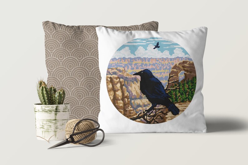 May include: A white pillow with a round, colorful image of a canyon landscape and a black raven perched on a branch. The raven is looking to the left. The image is surrounded by a white circle.