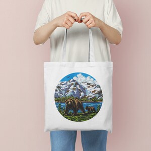 Pu&ograve; includere: Borsa tote in tela bianca con un design circolare che presenta un orso bruno e un cucciolo che camminano in un paesaggio montano con un lago blu e un cielo blu con nuvole bianche.