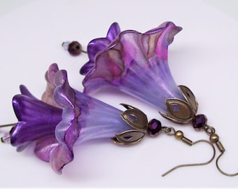 May include: A pair of handcrafted earrings featuring purple, bell-shaped flower pendants. The earrings have a metallic finish with bronze-colored accents and small, dangling beads. The floral design is detailed and elegant.