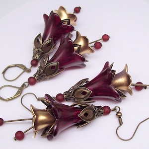May include: Earrings featuring burgundy and gold flower-shaped pendants with intricate metalwork. Each earring has a lever-back clasp and a stem with small red beads. The earrings are designed to resemble stylized flowers.