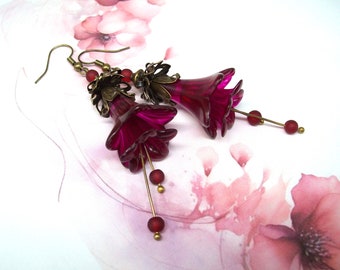 May include: A pair of dangle earrings featuring deep magenta flower-shaped pendants. The earrings have bronze-colored accents, including the hooks and decorative caps. Each earring has a slender gold-colored rod with red beads.