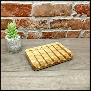 May include: A wooden soap dish with five slots for bar soap. The dish is made of light brown bamboo and has a natural finish. It is sitting on a wooden surface.
