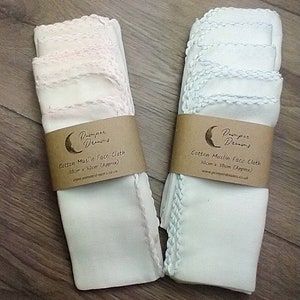 May include: Two stacks of white cotton muslin face cloths, each wrapped with a brown paper band. The cloths are approximately 12 inches by 12 inches, with a decorative stitched edge. The paper band reads "Pamper Dreams" and "Cotton Muslin Face Cloth".