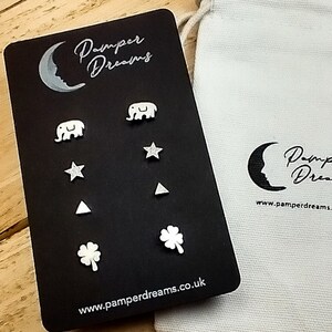 May include: A set of six silver stud earrings featuring various designs, including stars, elephants, triangles, and a four-leaf clover. The earrings are presented on a black card with the text "Pamper Dreams" and a website address. The earrings are packaged in a white drawstring bag with the same logo and website address.