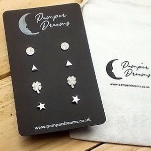 May include: A black card with the text "Pamper Dreams" and a crescent moon design. The card features six silver stud earrings with different designs: a circle, a triangle, a four-leaf clover, and a star. The card also includes the website address "www.pamperdreams.co.uk".