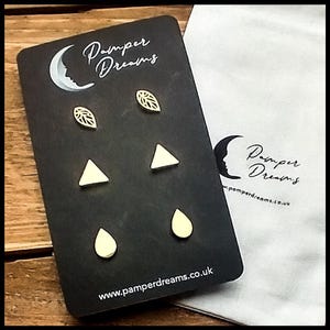 May include: A set of three pairs of earrings on a black card with the text "Pamper Dreams" and the website address "www.pamperdreams.co.uk". The earrings are made of wood and are in the shapes of leaves, triangles, and teardrops. The earrings are a light cream color.