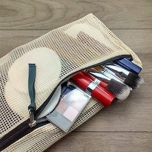 May include: A beige mesh makeup bag with a zipper closure. The bag is open and contains several makeup brushes, a red lipstick, and a compact eyeshadow palette.