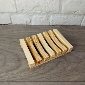 May include: A wooden soap dish with a slatted design. The dish is made of light-colored wood with a natural finish. The slats are arranged in a parallel pattern, allowing for drainage. The soap dish is designed to hold a bar of soap.