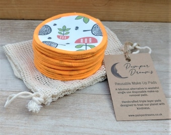 May include: A stack of 12 orange and white reusable makeup remover pads with a floral pattern. The pads are in a white mesh bag and next to a brown card with the text "Pamper Dreams Reusable Make Up Pads" and a website address.