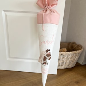Op de afbeelding: Een roze en witte schoolkegel met een paardenontwerp en de naam "Sarah" erop geborduurd. De kegel is versierd met witte stippen en heeft een roze lint aan de bovenkant.