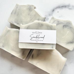 May include: Four bars of handmade sandalwood soap. The soap is white with gray swirls and has a label that reads "Kechi Bath Company Sandalwood Handcrafted Soap 5oz".