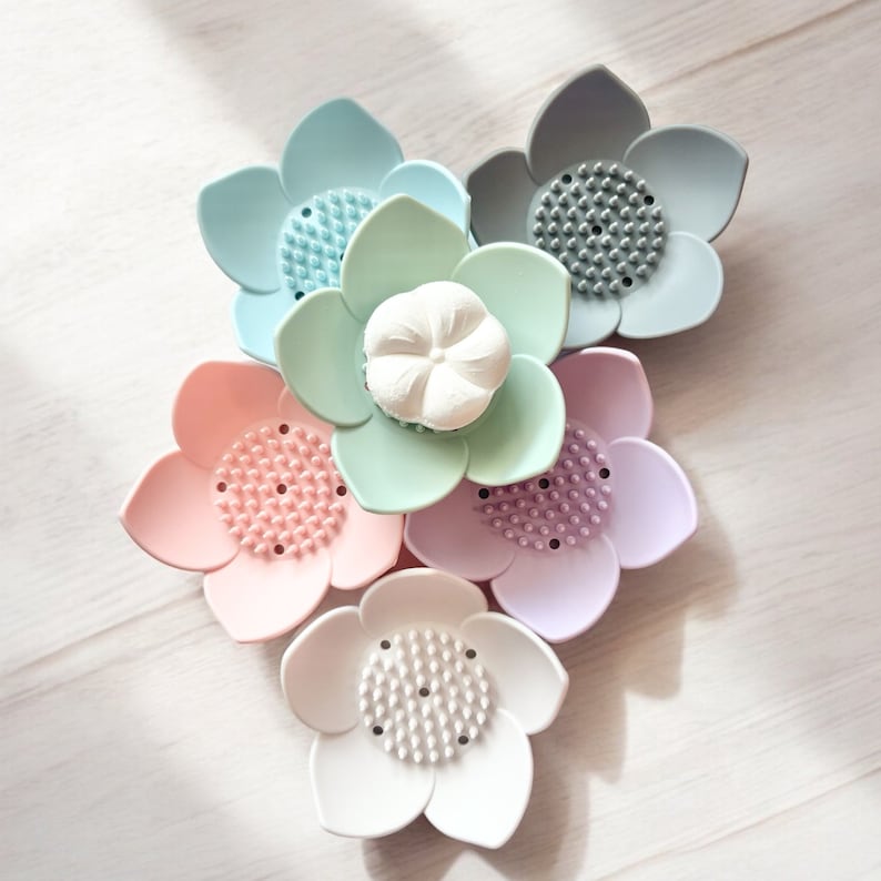 May include: Five pastel-colored silicone flower-shaped soap dishes. The dishes have a textured surface and are arranged in a circle. One dish has a white soap bar in the center.