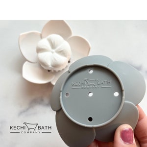 May include: Gray silicone bath flower with five petals and five holes. The center of the flower has the text "KECHI BATH COMPANY" with a small bathtub icon.