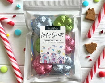 May include: A clear bag filled with foil-wrapped shower steamers, candy, and gingerbread cookies. The label reads "Land of Sweets" and lists the scents: Sugar Plum Fairy, Candy Cane, and Snickerdoodle. Candy canes and colorful candies surround the bag.